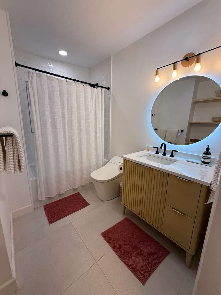 Clean, modern bathroom with a backlit round mirror, fluted wood vanity, white subway-tile shower and fresh towels